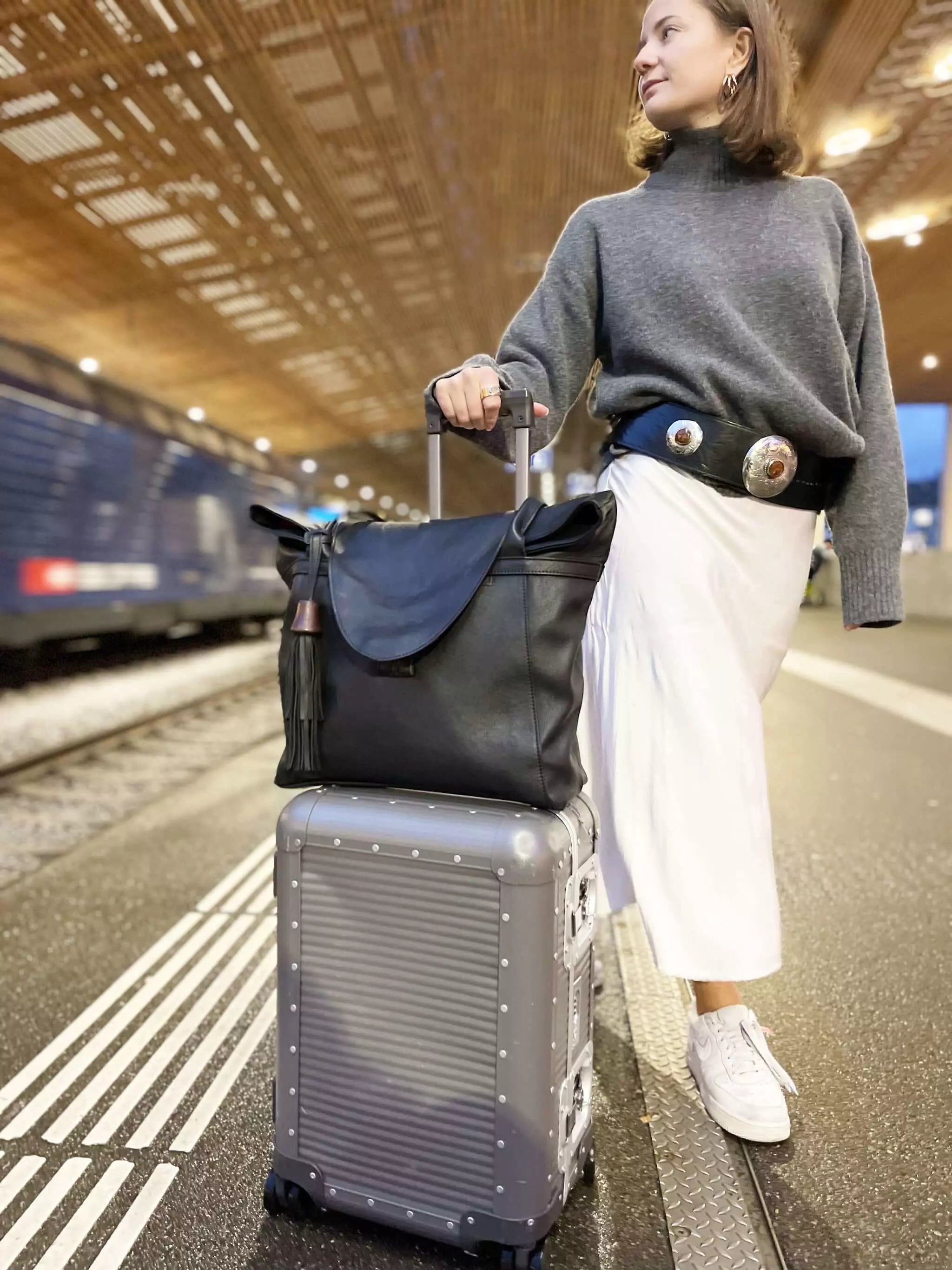 Frau trägt schwarze ABURY Shopper-Tasche auf einem Koffer und schwarzen Ledergürtel mit Metallapplikationen