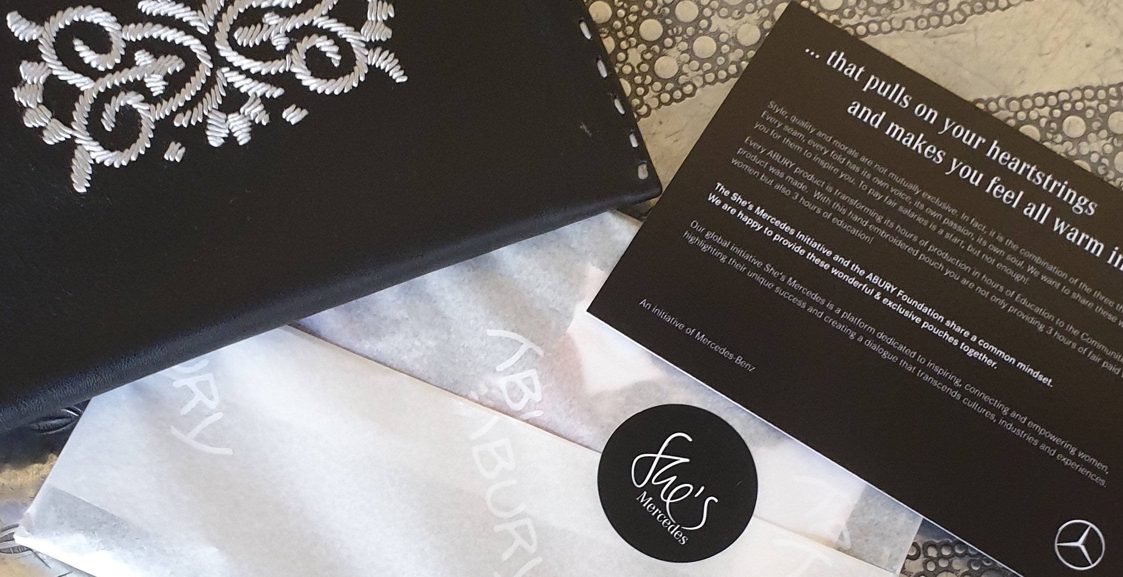 Schwarzes Ledertäschchen mit besticktem Blumenmotiv neben einem Mercedes-Flyer