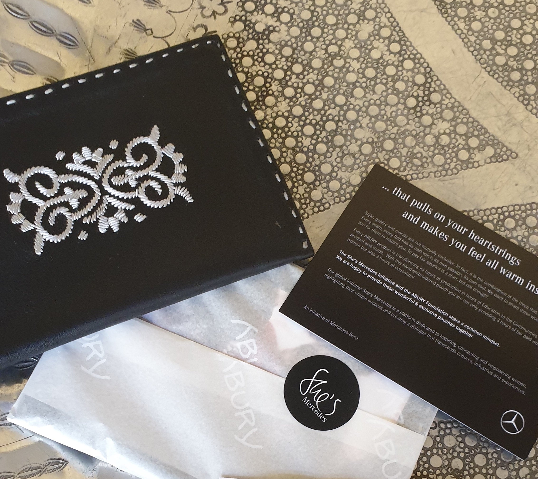 Schwarzes Ledertäschchen mit besticktem Blumenmotiv neben einem Mercedes-Flyer
