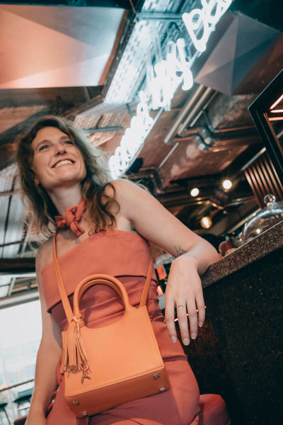 Die Frau sitzt auf einem Barhocker und trägt eine orangefarbene ABURY Mini Tote Bag. #farbe_orange