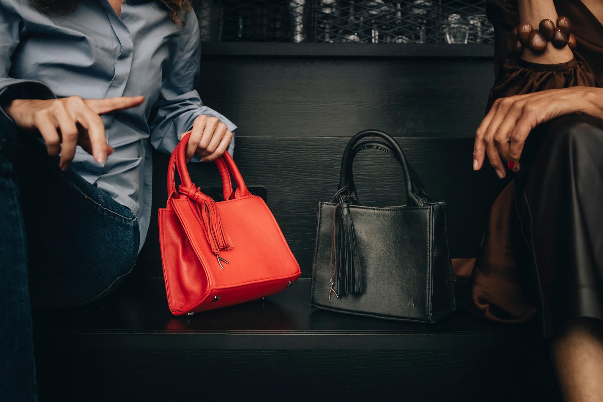 Zwischen zwei Frauen stehen eine rote und eine schwarze ABURY Mini Tote Bag. #farbe_schwarz