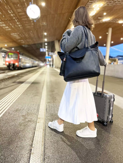 Frau trägt schwarze ABURY Shopper-Tasche auf einem Koffer und schwarzen Ledergürtel mit Metallapplikationen #farbe_schwarz