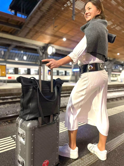 Frau trägt schwarze ABURY Shopper-Tasche auf einem Koffer und schwarzen Ledergürtel mit Metallapplikationen #farbe_schwarz