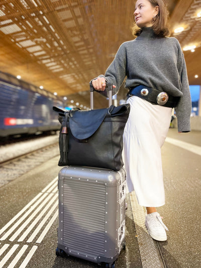 Frau trägt einen schwarzen Ledergürtel mit Metallapplikationen, neben ihr steht auf einem Koffer die schwarze Shopper-Tasche aus Leder. 