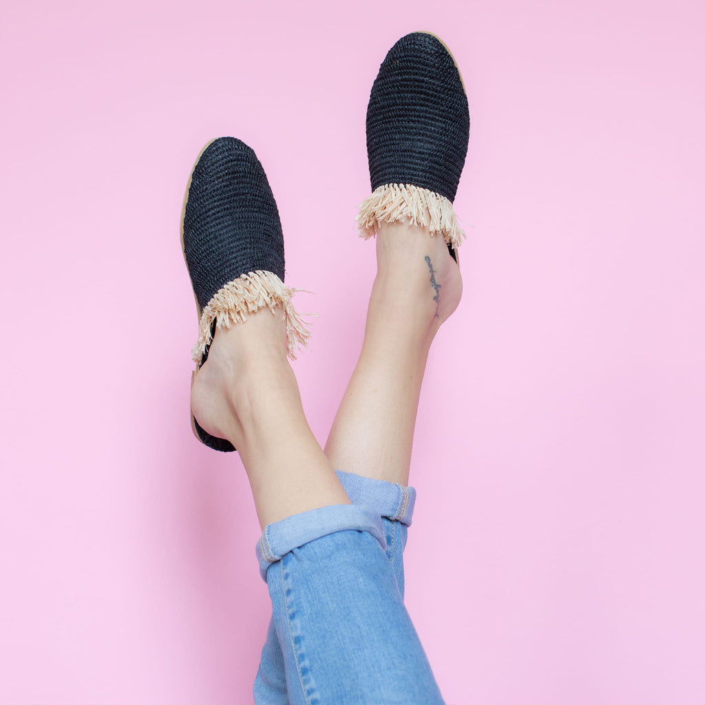 feet model in front of a pink background wearing jeans and abury black raffia summer slippers with fringes